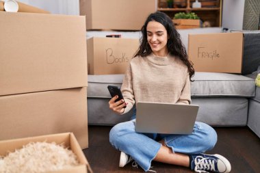 Young hispanic woman using laptop and smartphone sitting on floor at new home
