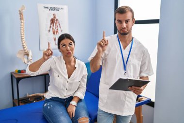Young hispanic woman at physiotherapist appointment pointing up looking sad and upset, indicating direction with fingers, unhappy and depressed. 