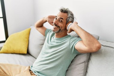 Orta yaşlı, kır saçlı, müzik dinleyen adam evdeki kanepede oturuyor.