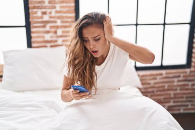 Genç, güzel, İspanyol bir kadın yatak odasında endişeli bir ifadeyle akıllı telefon kullanıyor.