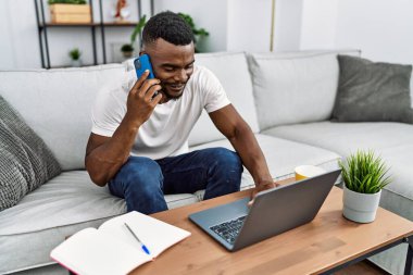 Young african american man using laptop talking on the smartphone at home