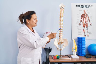 Orta yaşlı İspanyol kadın fizyoterapist rehabilitasyon kliniğinde omurganın anatomik modelini gösteriyor.