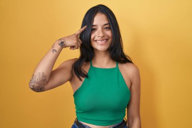 Brunette woman standing over yellow background smiling pointing to head with one finger, great idea or thought, good memory 