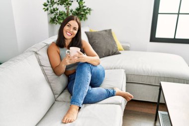 Genç ve güzel İspanyol kadın evdeki kanepede oturup kahve içiyor.