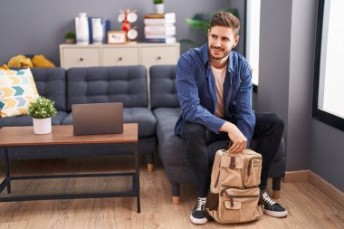 Beyaz bir adam sırt çantasını açıyor. Evdeki kanepede oturuyor.