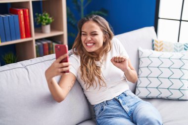 Young hispanic woman doing video call with smartphone screaming proud, celebrating victory and success very excited with raised arm 
