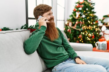 Young redhead man talking on the smartphone sitting by christmas tree at home