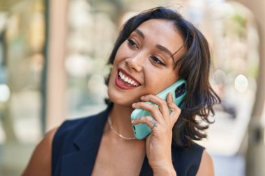 Genç, güzel, İspanyol bir kadın gülümsüyor ve caddede akıllı telefondan konuşuyor.