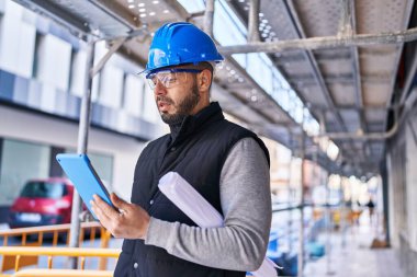 Young latin man architect using touchpad holding blueprints at street
