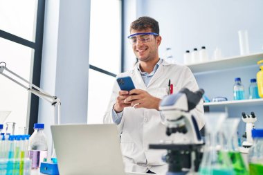 Young hispanic man scientist smiling confident using smartphone at laboratory