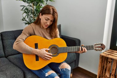 Orta yaşlı beyaz kadın klasik gitar çalıyor. Evdeki kanepede oturuyor..