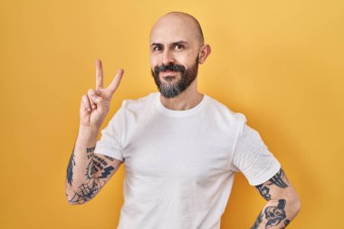 Sarı arka planda dikilmiş, gülümseyen ve parmaklarını zafer işareti yaparken gösteren İspanyol dövmeli genç bir adam. İki numara.. 