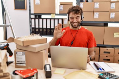 Young hispanic call center agent man working at warehouse waiving saying hello happy and smiling, friendly welcome gesture 