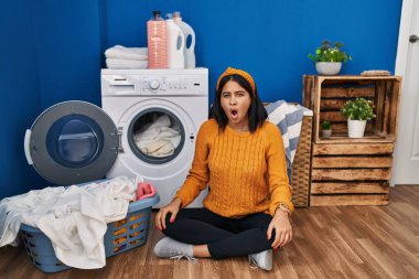 Young hispanic woman doing laundry in shock face, looking skeptical and sarcastic, surprised with open mouth 
