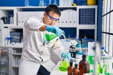 Young chinese man wearing scientist uniform measuring liquid at laboratory