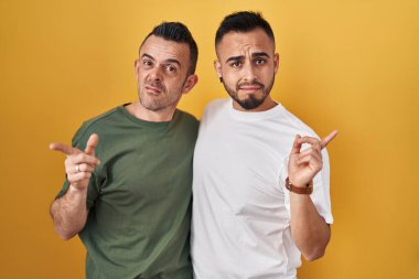 Homosexual couple standing over yellow background pointing aside worried and nervous with forefinger, concerned and surprised expression 