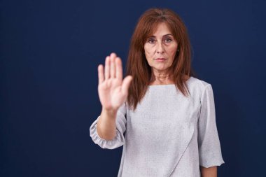 Middle age woman standing over blue background doing stop sing with palm of the hand. warning expression with negative and serious gesture on the face. 