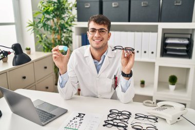Young hispanic oculist holding glasses and contact lenses smiling with a happy and cool smile on face. showing teeth. 