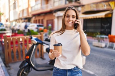 Young blonde woman talking on the smartphone and drinking coffee at street