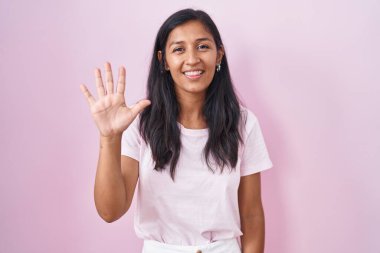 Young hispanic woman standing over pink background showing and pointing up with fingers number five while smiling confident and happy. 
