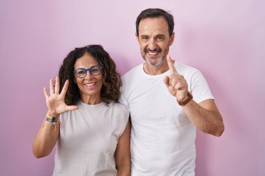 Orta yaşlı İspanyol bir çift pembe arka planda 6 numaralı parmaklarıyla işaret ederken kendinden emin ve mutlu bir şekilde gülümsüyor.. 