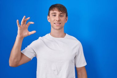 Caucasian blond man standing over blue background smiling and confident gesturing with hand doing small size sign with fingers looking and the camera. measure concept. 