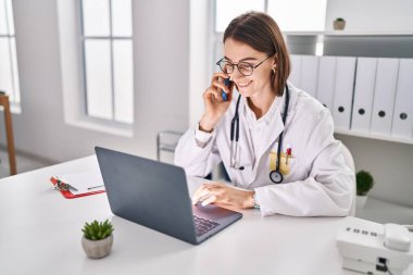 Young caucasian woman doctor using laptop talking on smartphone at clinic