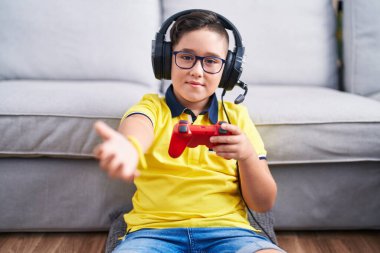 Young hispanic kid playing video game holding controller wearing headphones smiling cheerful offering palm hand giving assistance and acceptance. 