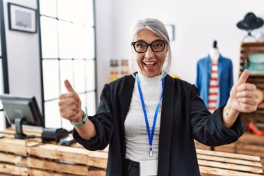 Orta yaşlı, gri saçlı, mağaza müdürü olarak çalışan, el hareketleriyle olumlu jest yapan, gülümseyen ve mutlu bir kadın. Neşeli ifade ve kazanan jest. 