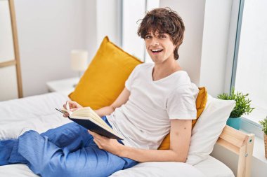 Young hispanic man reading book sitting on bed at bedroom