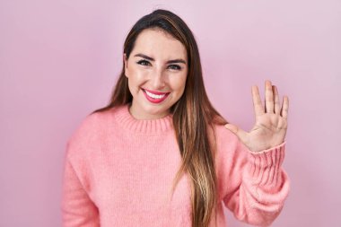 Young hispanic woman standing over pink background showing and pointing up with fingers number five while smiling confident and happy. 
