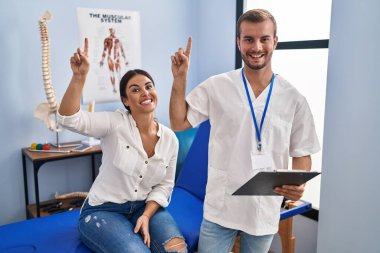 Young hispanic woman at physiotherapist appointment smiling amazed and surprised and pointing up with fingers and raised arms. 