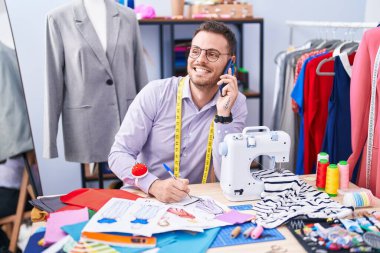 Genç İspanyol terzi terzide akıllı telefondan konuşuyor. Terzide defter üzerinde yazıyor.