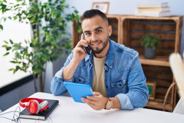 Young hispanic man using touchpad and talking on smartphone at home