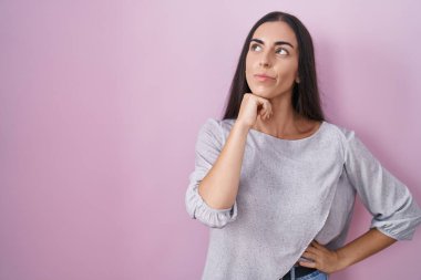 Pembe arka planda eli çenesinde duran genç esmer kadın soru sormayı, düşünceli yüz ifadesini düşünüyor. Düşünceli bir yüzle gülümsüyor. şüphe kavramı. 