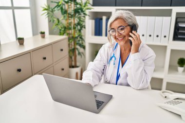 Klinikte doktor üniforması giyen orta yaşlı bir kadın akıllı telefondan konuşuyor.