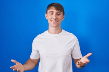Caucasian blond man standing over blue background smiling cheerful with open arms as friendly welcome, positive and confident greetings 