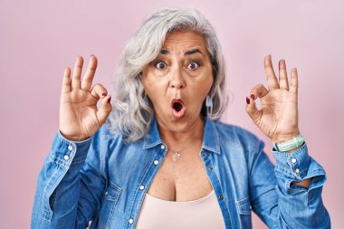 Pembe arka planda duran gri saçlı orta yaşlı kadın parmaklarıyla onay sembolü yaparken şaşırmış ve şok olmuş görünüyor. çılgın ifade 