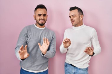 Homosexual couple standing over pink background disgusted expression, displeased and fearful doing disgust face because aversion reaction. 
