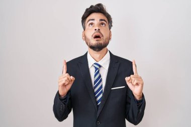 Young hispanic man with tattoos wearing business suit and tie amazed and surprised looking up and pointing with fingers and raised arms. 