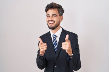 Young hispanic man with tattoos wearing business suit and tie pointing fingers to camera with happy and funny face. good energy and vibes. 
