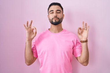 İspanyol genç adam pembe arka planda duruyor rahatlamış ve gözleri kapalı gülümsüyor parmaklarıyla meditasyon hareketi yapıyor. yoga konsepti. 