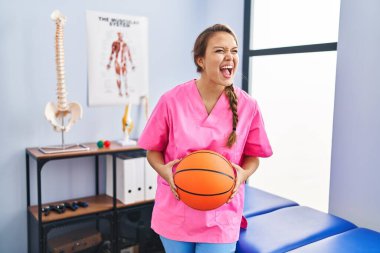 Young hispanic woman working at physiotherapy clinic holding basketball ball angry and mad screaming frustrated and furious, shouting with anger. rage and aggressive concept. 