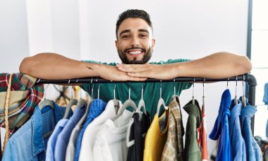 Genç Arap müşteri gülümsüyor giyim mağazasında askılara yaslanıyor.