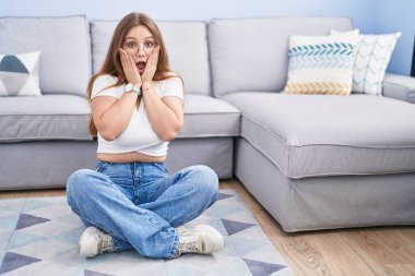 Oturma odasında yerde oturan genç beyaz kadın korkmuş ve şok olmuş, şaşırmış ve şaşırmış bir halde. 