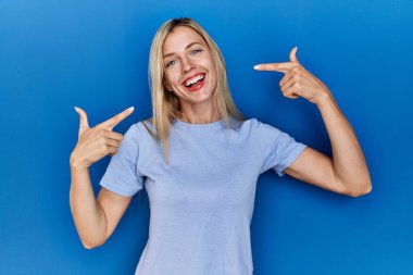 Beautiful blonde woman wearing casual t shirt over blue background smiling cheerful showing and pointing with fingers teeth and mouth. dental health concept. 