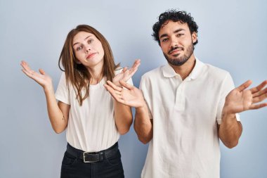 Sıradan kıyafetler giyen genç bir çift, elleri ve kolları havada, budala ve kafası karışmış bir şekilde yan yana duruyorlar. şüphe kavramı. 