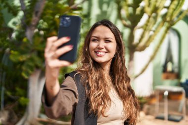 Genç, güzel, İspanyol bir kadın gülümsüyor. Parktaki akıllı telefonun yanında özçekim yapıyor.