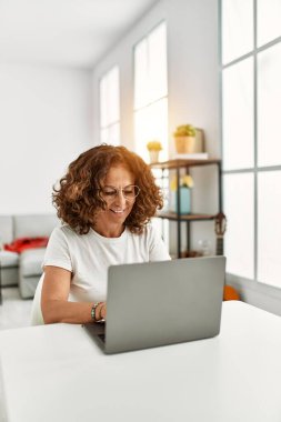 Orta yaşlı İspanyol kadın evde dizüstü bilgisayar kullanarak gülümsüyor.