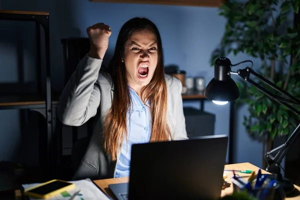Young brunette woman working at the office at night angry and mad raising fist frustrated and furious while shouting with anger. rage and aggressive concept.